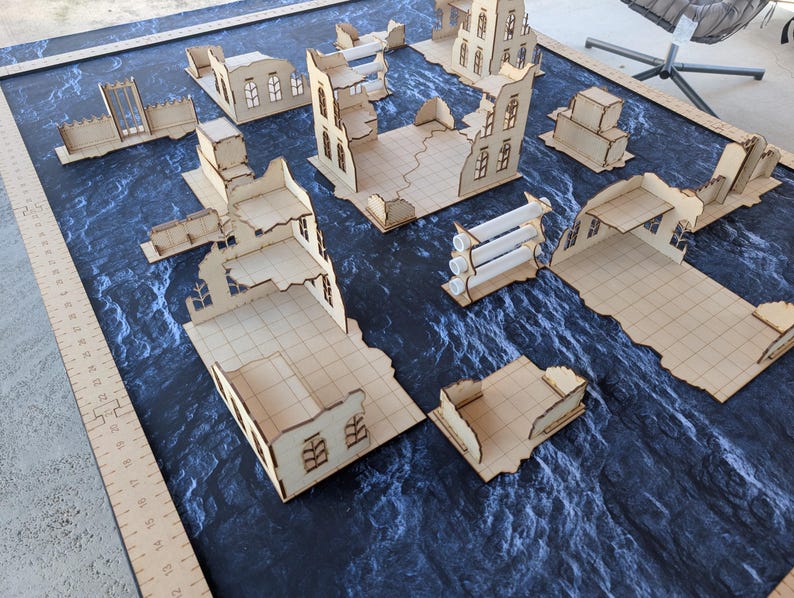 May include: A collection of laser-cut light wood miniature building ruins for tabletop gaming, arranged on a dark blue textured mat. A wooden ruler along the left edge shows measurements in inches, with structures featuring grid floors and walls.