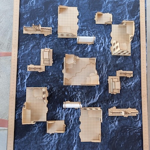 May include: A set of laser-cut wooden tabletop wargaming terrain pieces featuring modular building ruins with grid-patterned floors, arranged on a dark textured mat framed by a wooden ruler measuring approximately 24 inches by 36 inches.