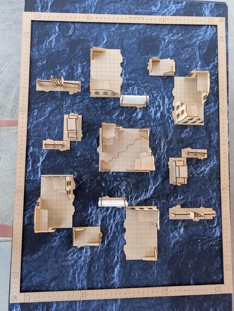 May include: A set of laser-cut wooden tabletop wargaming terrain pieces featuring modular building ruins with grid-patterned floors, arranged on a dark textured mat framed by a wooden ruler measuring approximately 24 inches by 36 inches.