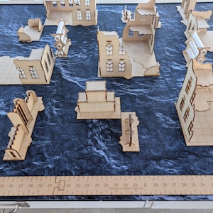 May include: A tabletop gaming terrain set featuring laser-cut light wood building ruins and walls arranged on a dark textured mat. A wooden ruler is visible along the bottom and left edges, showing measurements in inches for scale.