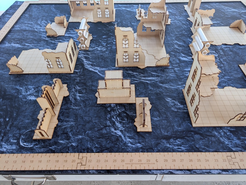 May include: A tabletop gaming terrain set featuring laser-cut light wood building ruins and walls arranged on a dark textured mat. A wooden ruler is visible along the bottom and left edges, showing measurements in inches for scale.