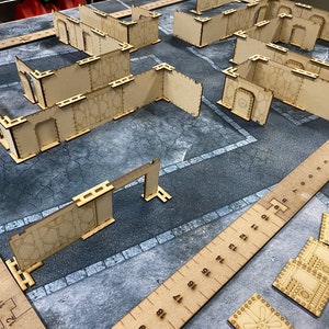 Puede incluir: Un conjunto de terreno para juegos de mesa hecho de madera marr&oacute;n claro. El conjunto incluye paredes, puertas y herramientas de medici&oacute;n, dispuestas sobre una superficie gris. Las paredes tienen cortes decorativos y est&aacute;n dise&ntilde;adas para crear un dise&ntilde;o de ciudad o edificio para juegos de mesa.