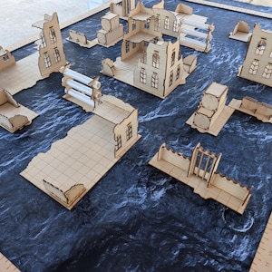 May include: A collection of laser-cut wooden miniature building ruins for tabletop gaming, featuring various wall sections, multi-story structures, and a small rack with three clear plastic tubes, arranged on a dark mat with visible inch rulers.