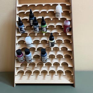 May include: A wooden paint bottle organizer with multiple shelves and curved cutouts to hold small bottles. The organizer is filled with various paint bottles in different colors, including black, white, green, and pink. The organizer is made of light brown wood.