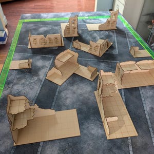 May include: A collection of laser-cut, unfinished wooden terrain pieces for tabletop gaming. The set includes various building ruins, walls, and platforms, arranged on a gray and green gaming mat. The pieces are designed to create a detailed battlefield.
