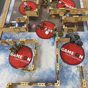 Puede incluir: Una escena de juego de mesa con figuras en miniatura y terreno. La disposici&oacute;n incluye estructuras de madera, alfombras circulares rojas con el texto "GAME ON CHATTANOOGA" y una superficie de juego con patrones azules y blancos. Las figuras en miniatura est&aacute;n posicionadas.