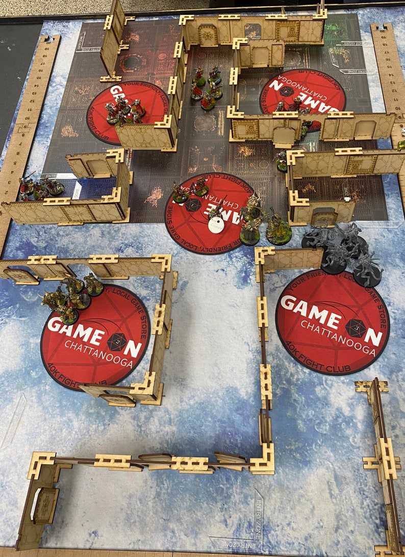 Puede incluir: Una escena de juego de mesa con figuras en miniatura y terreno. La disposici&oacute;n incluye estructuras de madera, alfombras circulares rojas con el texto "GAME ON CHATTANOOGA" y una superficie de juego con patrones azules y blancos. Las figuras en miniatura est&aacute;n posicionadas.