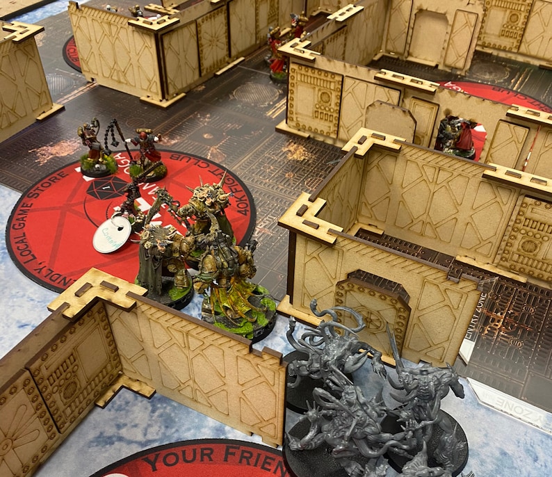 Puede incluir: Una escena de juego de mesa con figuras en miniatura y terreno modular. Las figuras est&aacute;n pintadas en varios colores y se colocan en un tablero de juego con un dise&ntilde;o circular rojo. El terreno incluye muros y estructuras cortadas con l&aacute;ser.