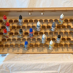 May include: A light brown wooden tiered display rack featuring multiple circular cutouts designed to hold small paint bottles. The rack is wall-mountable with two keyhole slots at the top and contains several assorted paint bottles with colored caps.