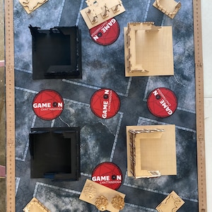May include: A tabletop role-playing game setup featuring a textured gray and black mat with various laser-cut wooden terrain pieces, including walls, buildings, and decorative elements. Several red circular logos with white text are scattered across the mat.