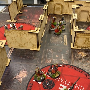 Puede incluir: Una escena de juego de guerra de mesa con figuras en miniatura y terreno modular. Las figuras, pintadas en varios colores, est&aacute;n colocadas en un tablero de juego detallado con detalles en rojo y gris. Las estructuras de madera crean un entorno laber&iacute;ntico.