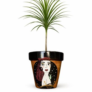 Può includere: Vaso di fiori decorativo in terracotta con un'illustrazione dipinta a mano di una donna con i capelli rossi e un foulard rosso. Il vaso contiene una pianta verde con foglie lunghe e sottili. Il vaso ha un bordo nero.