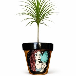 Può includere: Vaso di fiori in terracotta con bordo nero, con un'illustrazione dipinta a mano di una donna dai capelli rossi che tiene un uccello blu. Il vaso contiene una pianta verde con foglie lunghe e sottili.