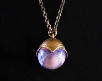 May include: A necklace featuring a round, marbled pendant with swirls of purple, pink, and white. The pendant is encased in a bronze-toned metal cap with a pointed, hood-like design and hangs from a matching bronze-toned metal chain link necklace.