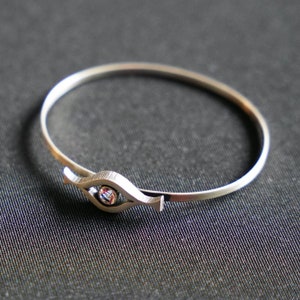 May include: A silver bangle bracelet with a single clear gemstone set in a stylized eye shape.