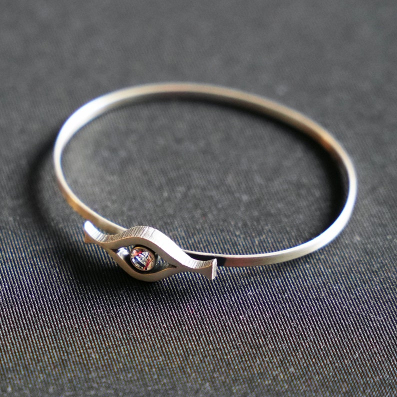 May include: A silver bangle bracelet with a single clear gemstone set in a stylized eye shape.