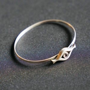 May include: A silver bangle bracelet with a unique eye-shaped design featuring a clear gemstone in the center.