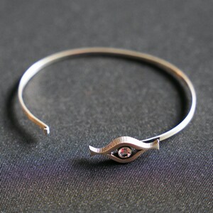 May include: A silver bangle bracelet with a single clear gemstone set in a stylized eye design.