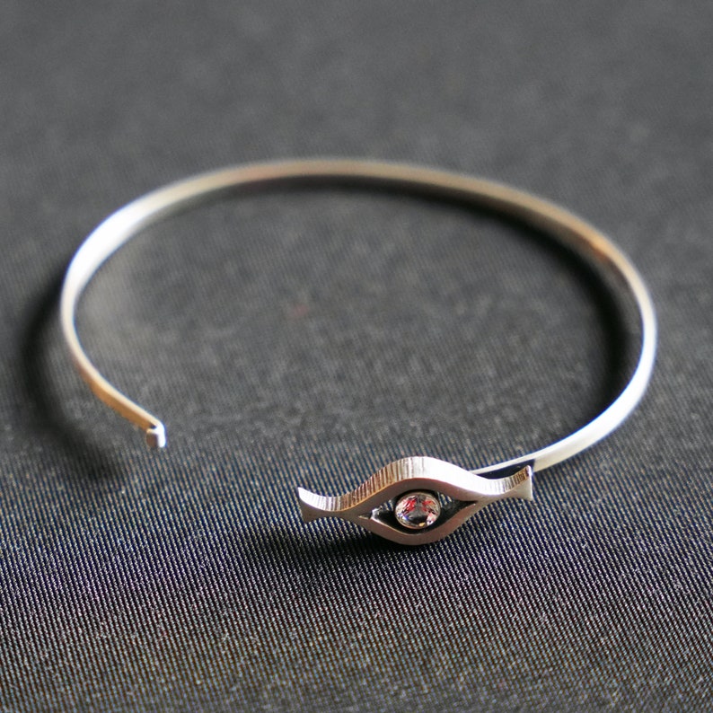 May include: A silver bangle bracelet with a single clear gemstone set in a stylized eye design.