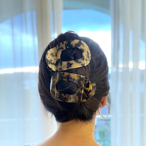 May include: Two oval-shaped hair clips in a tortoiseshell pattern are positioned in the hair. The clips are a mix of cream, brown, and black. The person is wearing a black top with a floral lace trim and gold hoop earrings.