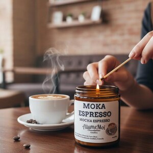 May include: A brown glass candle jar with a white label reading MOKA ESPRESSO and AllumeMoi, positioned next to a white ceramic cup of coffee with latte art and steam rising, while a match is used to light the candle wick on the table.