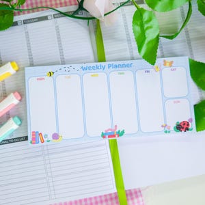 Könnte beinhalten: Ein hellblauer Wochenplaner mit dem Text "Weekly Planner" und den Wochentagen. Der Planer hat Cartoon-Illustrationen einer Biene, eines Schmetterlings, eines Marienkäfers und anderer kleiner Kreaturen. Drei bunte Stifte sind im Bild zu sehen.