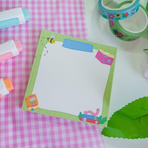 Op de afbeelding: Een vierkant, blanco notitieblok met een groene rand en decoratieve illustraties van een bij, een worm en boeken. Het notitieblok ligt op een roze en witte geruite ondergrond, met kleurrijke plakbandrollen en gummen in de buurt.