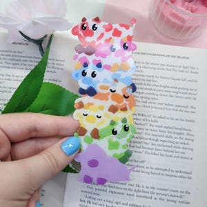 May include: A vertical bookmark featuring a stack of seven cartoon cows in a rainbow gradient of colours including red, pink, blue, orange, yellow, green, and purple. Each cow has a distinct expression and spots, creating a colourful, layered design.