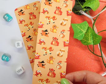 May include: Two rectangular bookmarks featuring a repeating pattern of various orange, red, and yellow cartoon creatures on a light orange background. The bookmarks are displayed alongside several multi-sided dice and green artificial leaves.