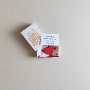 May include: A small white box with a pink and white gift box design on the lid. The box is open and contains a small red wooden heart and a card that reads "A little Heart From me to you Just to say I miss you!"