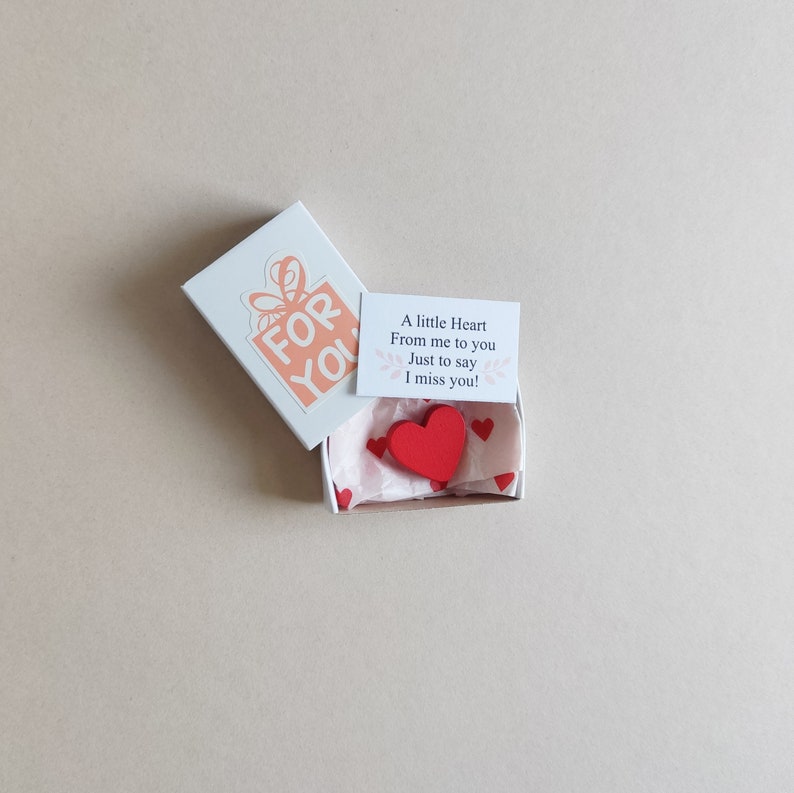 May include: A small white box with a pink and white gift box design on the lid. The box is open and contains a small red wooden heart and a card that reads "A little Heart From me to you Just to say I miss you!"