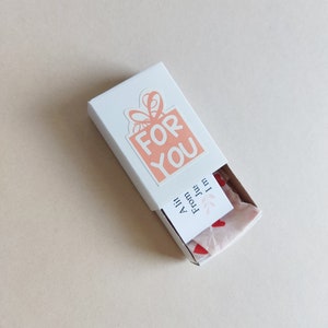 May include: A white gift box with a pink and white sticker that says "FOR YOU". The box is open and there is pink tissue paper with red hearts inside. The tissue paper has a small white tag that says "From Julie" and "All the best!"