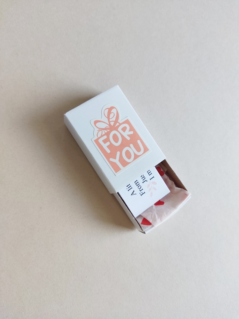 May include: A white gift box with a pink and white sticker that says "FOR YOU". The box is open and there is pink tissue paper with red hearts inside. The tissue paper has a small white tag that says "From Julie" and "All the best!"
