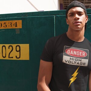 May include: A man wearing a black t-shirt with a yellow lightning bolt and a red and white sign that says "Danger High Voltage".