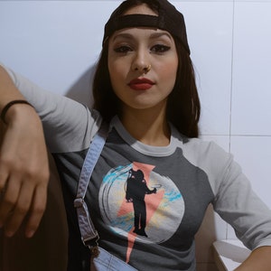 Puede incluir: Una camiseta de béisbol gris y negra con un diseño gráfico que presenta la silueta de una persona con una mochila y un rayo. La persona lleva una gorra de béisbol negra.