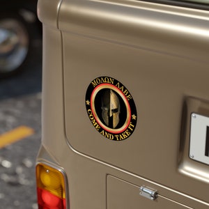 May include: A round sticker with a Spartan helmet design. The sticker has the text "MOLON LABE" at the top and "COME AND TAKE IT" at the bottom, with stars on each side. The helmet is gold, and the text is black.