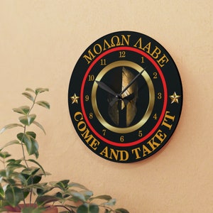 May include: Round black wall clock with gold Roman numerals and hands. The clock features a gold Spartan helmet design in the center and the phrases "MOLON LABE" and "COME AND TAKE IT" in gold lettering. Two gold stars are on the clock face.