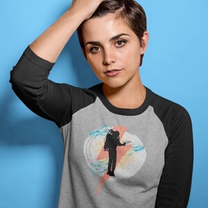 Op de afbeelding: Een grijs raglan baseballshirt met zwarte mouwen. Het shirt heeft een afbeelding van een duiker silhouet in een witte cirkel met een roze bliksemschicht en blauwe en gele accenten.