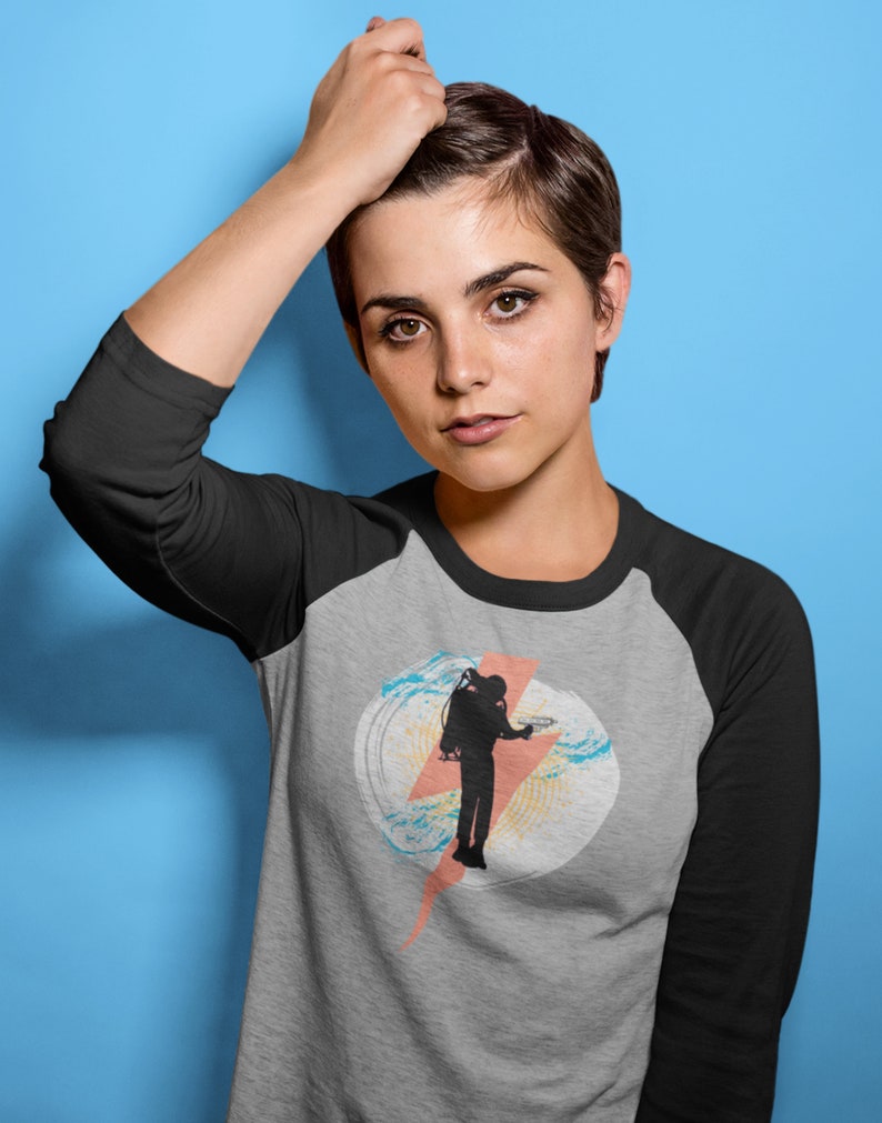 Op de afbeelding: Een grijs raglan baseballshirt met zwarte mouwen. Het shirt heeft een afbeelding van een duiker silhouet in een witte cirkel met een roze bliksemschicht en blauwe en gele accenten.