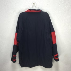 May include: A navy blue jacket with red accents on the sleeves and collar, hanging on a white hanger. The jacket has a full zipper and a high collar. The back of the jacket is visible.
