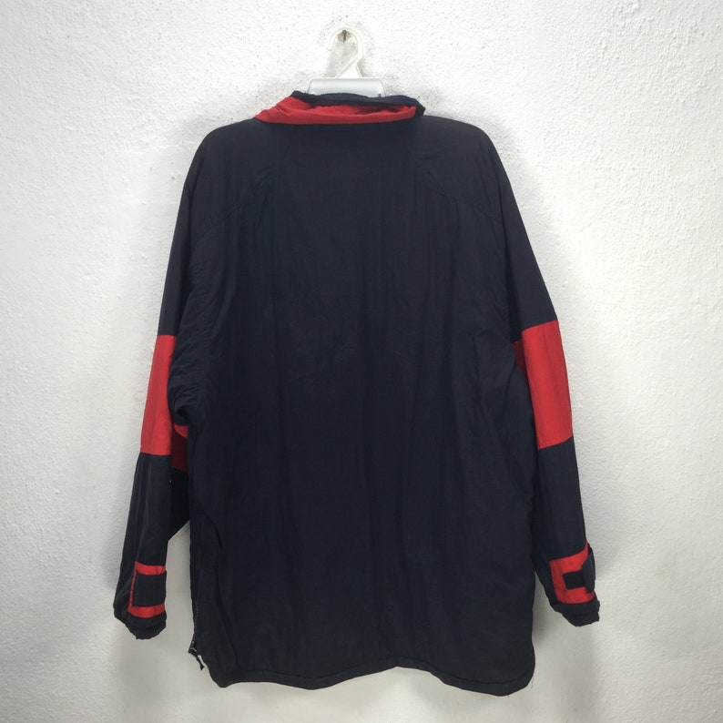 May include: A navy blue jacket with red accents on the sleeves and collar, hanging on a white hanger. The jacket has a full zipper and a high collar. The back of the jacket is visible.