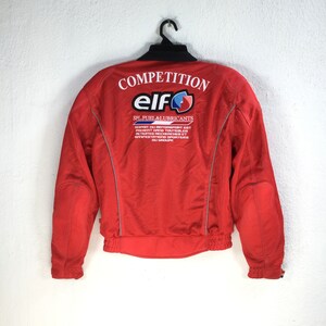 May include: Red racing jacket with a white "COMPETITION" logo and a white "elf" logo with a blue and red graphic. The text "SPL FUEL & LUBRICANTS" is also printed in white.