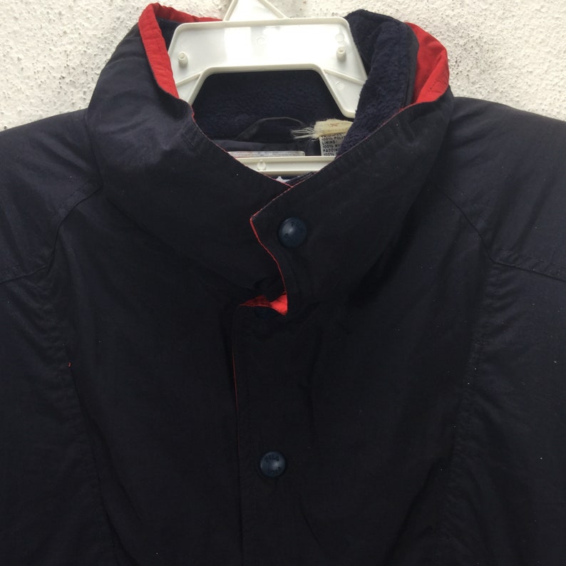 May include: Close-up of a navy blue jacket with a red collar lining and two navy blue buttons. The jacket is hanging on a white hanger. The collar is a high, stand-up style.