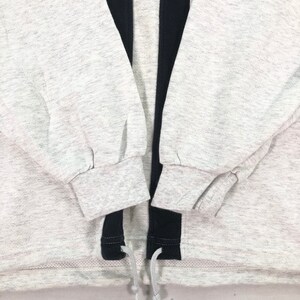 May include: A light gray sweatshirt with black accents and a drawstring hem. The sweatshirt features a small red logo on the upper left chest and black mesh detailing on the shoulders. The sleeves are cuffed at the wrists.