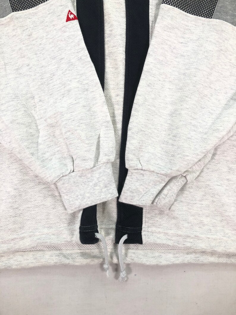 May include: A light gray sweatshirt with black accents and a drawstring hem. The sweatshirt features a small red logo on the upper left chest and black mesh detailing on the shoulders. The sleeves are cuffed at the wrists.