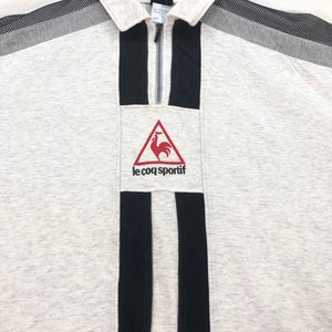 May include: A light gray sweatshirt with a collared neckline and a quarter-zip closure. The shirt features black vertical stripes and a red rooster logo with the text "le coq sportif". The shoulders have gray and black mesh accents.