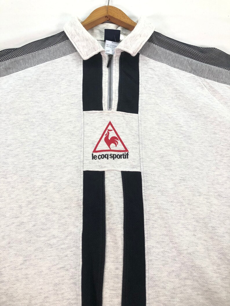 May include: A light gray sweatshirt with a collared neckline and a quarter-zip closure. The shirt features black vertical stripes and a red rooster logo with the text "le coq sportif". The shoulders have gray and black mesh accents.