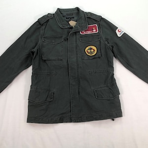 Może przedstawiać: Zielona kurtka typu utility z wieloma kieszeniami i zamkiem błyskawicznym. Kurtka ma kilka naszywek, w tym jedną z napisem "The Salty Siren".