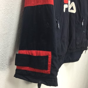May include: A navy blue jacket with red accents and the Fila logo in white. The jacket features a red panel across the shoulders and a red-trimmed pocket. Zippers are visible on the front.