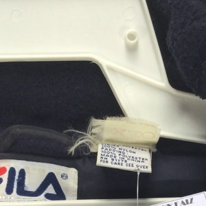 May include: Close-up of a navy blue and red FILA jacket. The jacket is hanging on a white hanger. The FILA logo is visible on the jacket. A tag with care instructions is also visible.